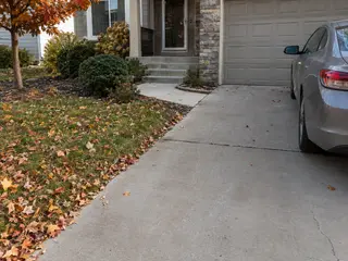 Driveway with minor wear beside a walkway, porch steps, and a parked car in autumn.